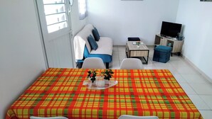 Un coin repas avec une table recouverte d’une nappe à carreaux, deux chaises blanches et une petite table sur laquelle est posé un vase de fleurs.
