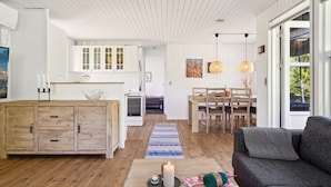 A spacious living area with a wooden floor, a dark gray sofa, a coffee table with a candle and knitting materials, a dining area with a wooden table and chairs, and a kitchen with white cabinets and a stove. - "Aldina" - 400m to the fjord by Interhome (Havndal)