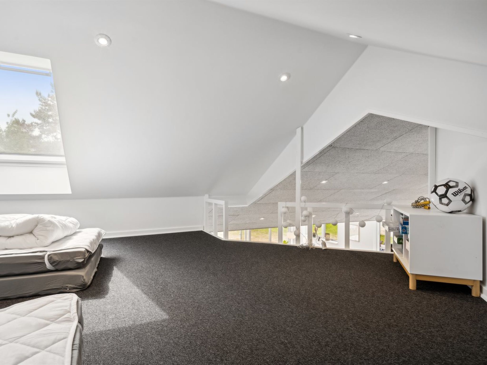 Une chambre moderne et bien éclairée, située sous les combles, avec un plafond en pente, une grande fenêtre et une vue sur l’extérieur.