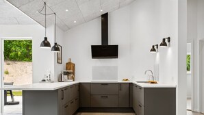 Una cocina moderna con una isla central, armarios gris oscuro, un televisor de pantalla plana montado sobre la isla, iluminación colgante y vistas a zonas verdes.