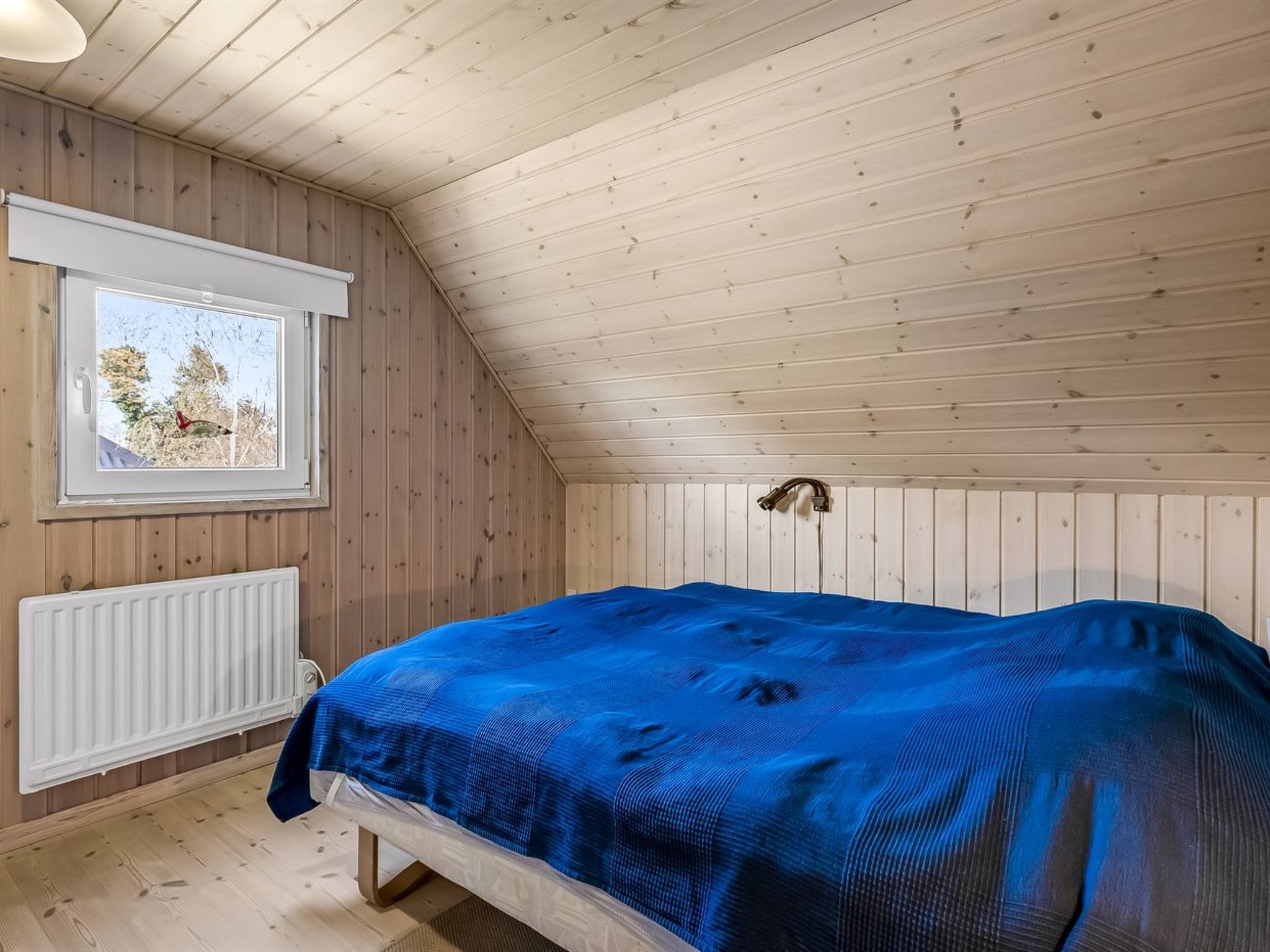 Un dormitorio con techo inclinado, una ventana con vistas a árboles, un radiador y una cama con cubrecama azul.