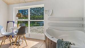 A modern bathroom with a large bathtub, a small table with chairs, and a window with a view of a wooden deck and trees.