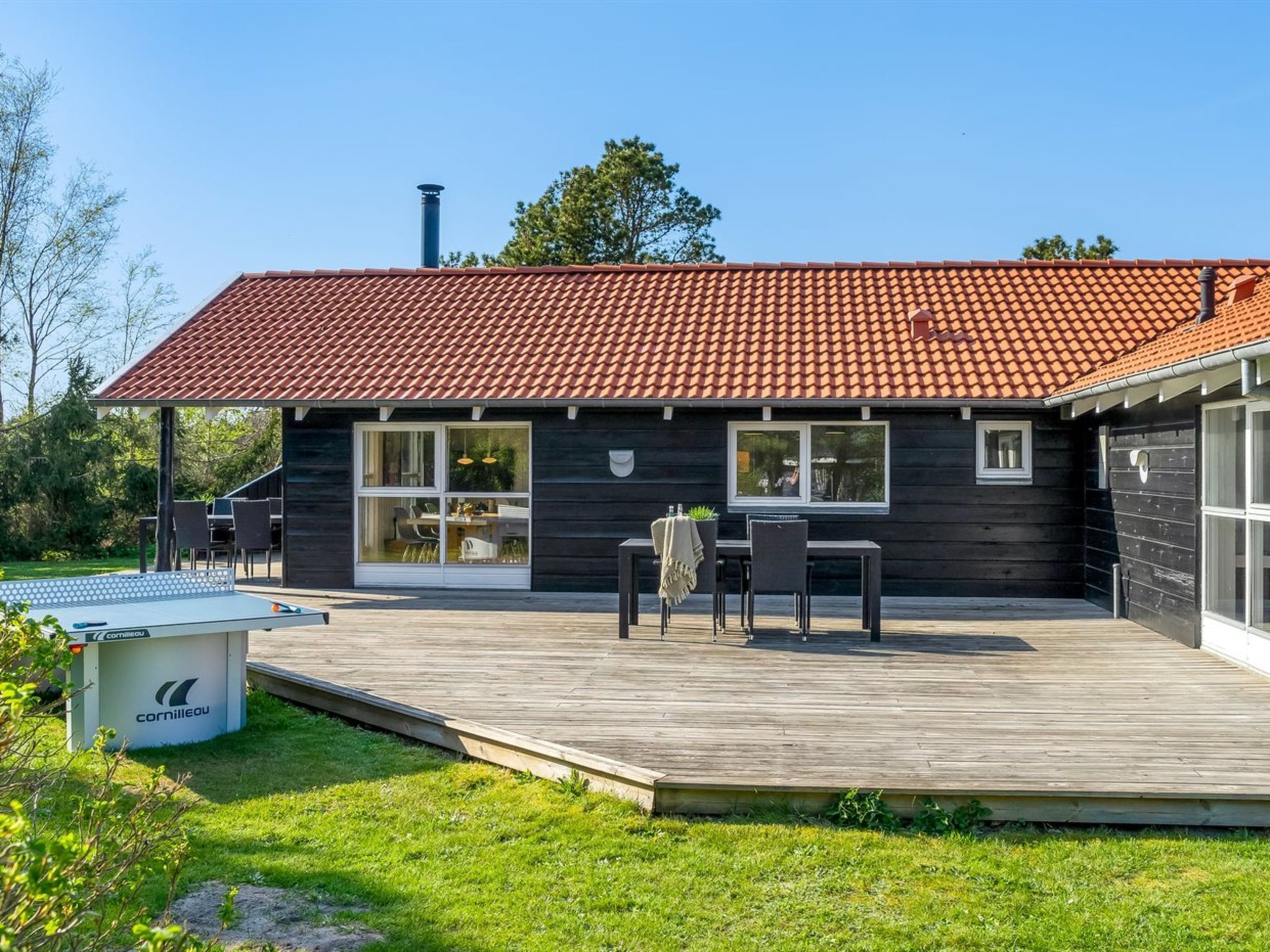 Una casa moderna con techo de tejas rojas, una terraza con mobiliario exterior y un amplio frente con ventanas.