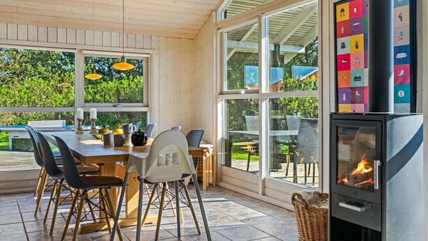 A cozy outdoor dining area with a wooden table and chairs, large windows, and a wood-burning stove.