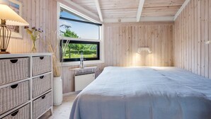 A room with wooden walls and ceiling, a bed with a white bedspread, a window with a view of greenery, a white dresser with wicker baskets, and a wall-mounted lamp. - "Ingwin" -  from the sea by Interhome (Hals)