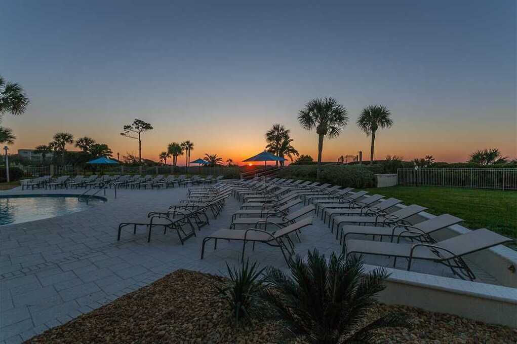 Un espace aménagé au bord de la piscine, avec des chaises longues et des parasols, des palmiers, et un coucher de soleil en arrière-plan.