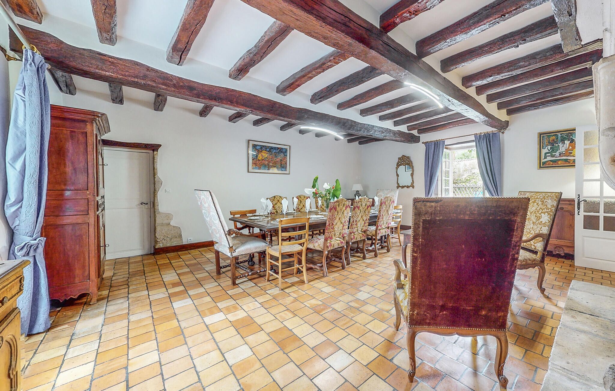 Un comedor con una amplia mesa de madera, sillas y una butaca de terciopelo rojo. Se observa una escalera con una puerta de madera y una ventana con cortinas.