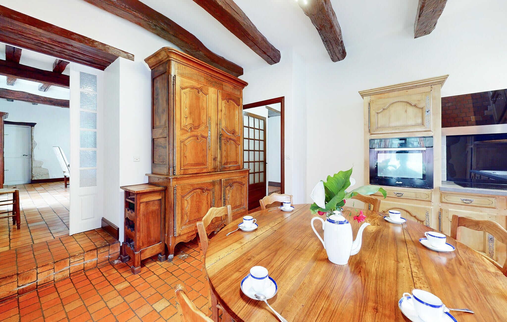 Une salle à manger avec une table en bois dressée pour quatre personnes, entourée de chaises. On aperçoit, au fond, une grande armoire en bois dotée de portes en verre.