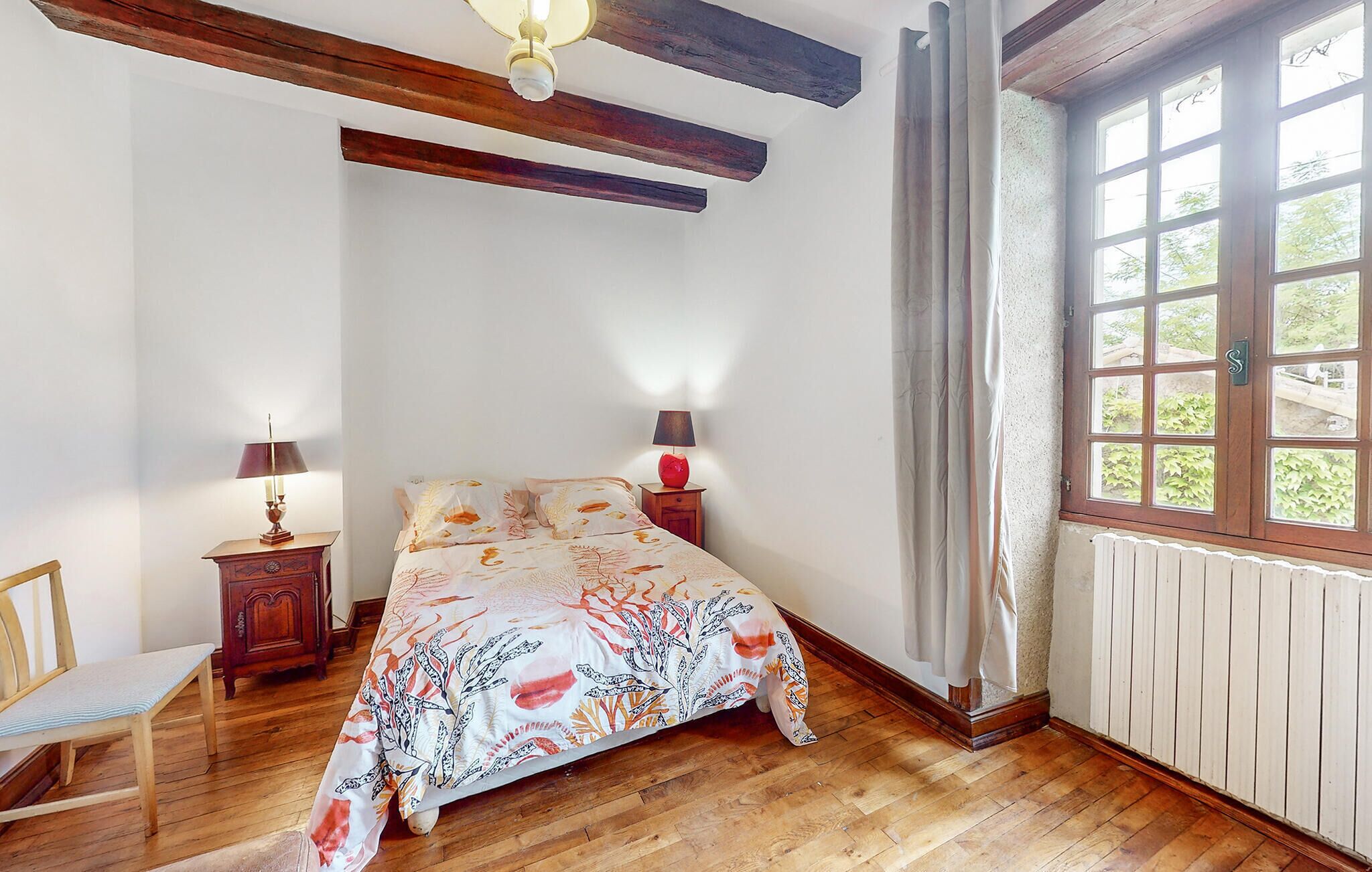 A bedroom with a wooden floor, a bed with a floral bedspread, two bedside tables with lamps, a chair, and a window with a view of greenery.