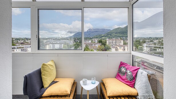 Un balcón con vistas a un paisaje urbano y montañas, que incluye una pequeña mesa con un vaso y una jarrón, y dos bancos con cojines.