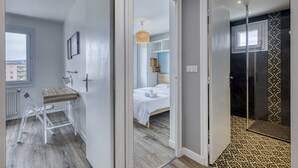 Una habitación de hotel moderna con un escritorio de madera, una cama con ropa blanca y una zona de ducha con azulejos estampados.