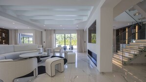 Un salon moderne avec un grand canapé d’angle, une table basse et un téléviseur à écran plat. Un escalier avec des rampes en verre mène à un niveau supérieur.
