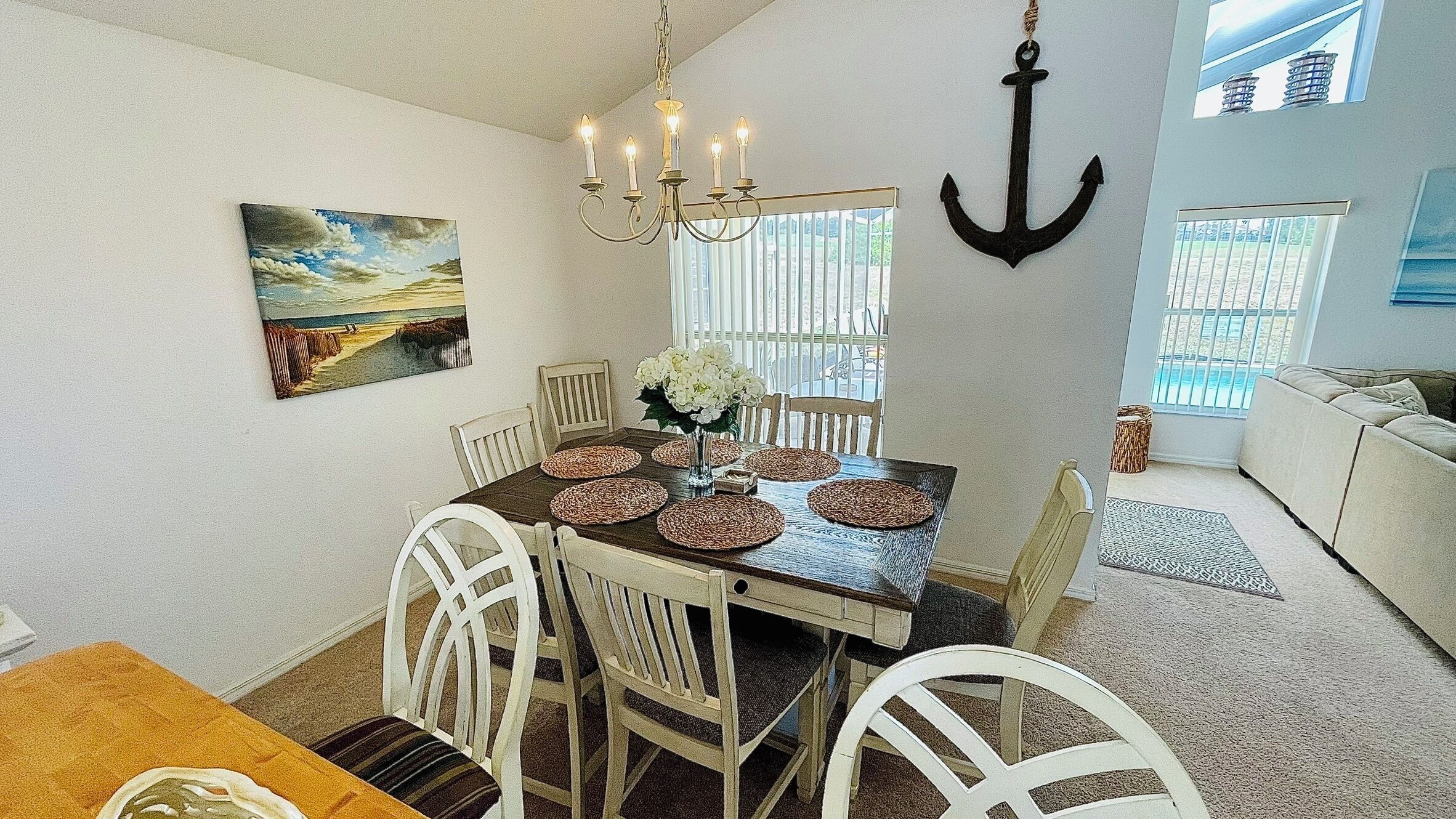 A dining area with a table set for six, surrounded by white chairs, a chandelier, and a painting of a beach scene.