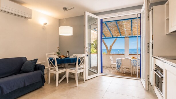 A modern kitchen with white cabinets, a dining area with a blue tablecloth, and a view of the sea through an open door. - Tra il Borgo e il Mare-Bel Horizonte - Tra il Borgo e il Mare - Bel Horizonte (Capoliveri)