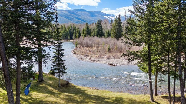 A riverside campsite with a fire pit and seating area, surrounded by trees and mountains. - Gods County Cottage on Swan River (Bigfork)