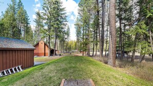 A rustic cabin with a wooden deck and a fire pit in the center, surrounded by tall pine trees. - Gods County Cottage on Swan River (Bigfork)
