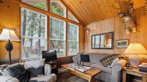 A cozy cabin interior with a wooden ceiling, large windows, a mounted deer head, a television, and a fireplace. - Gods County Cottage on Swan River (Bigfork)