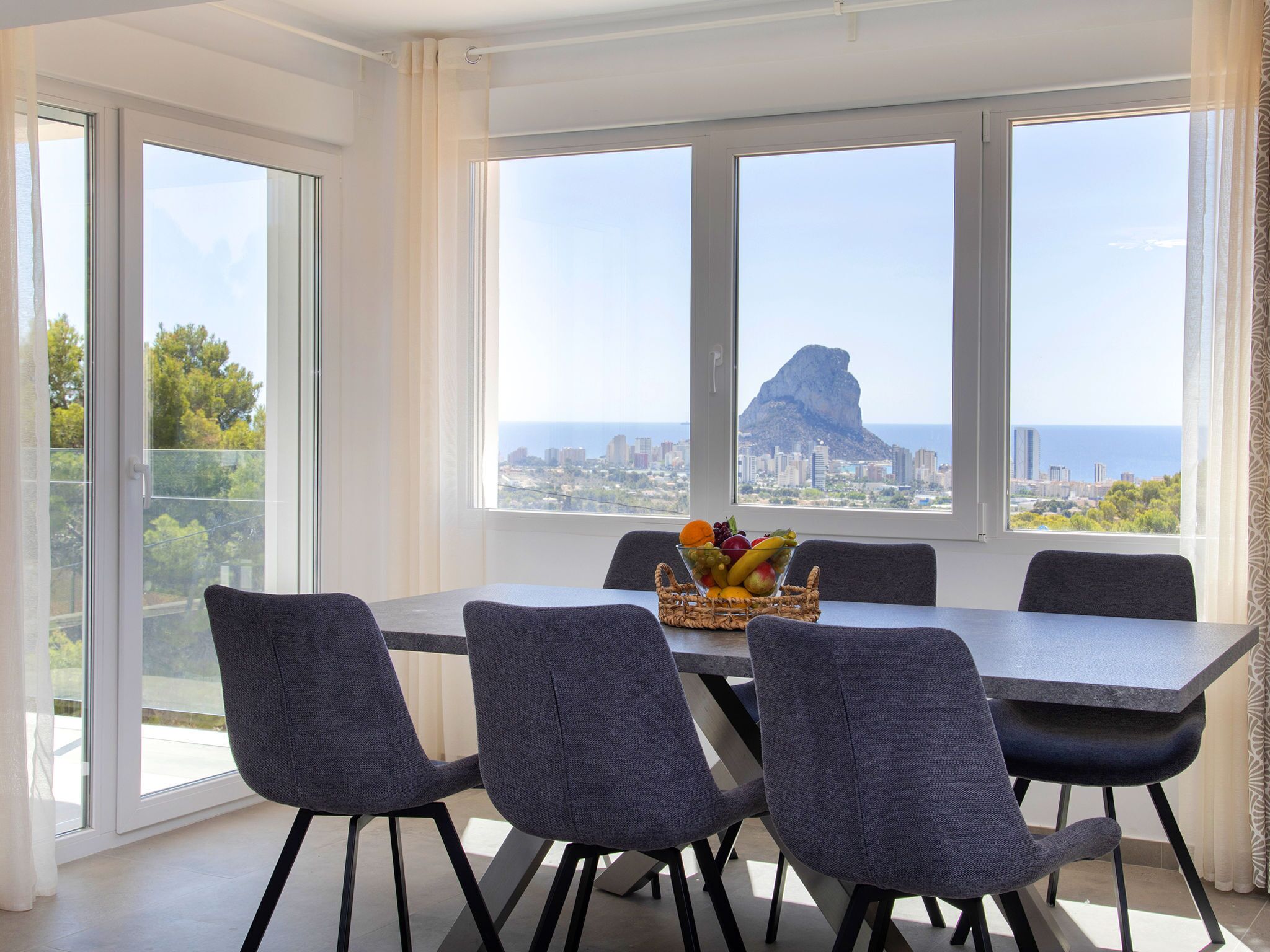 Un comedor con una mesa y sillas, un frutero y vistas a la ciudad a través de amplias ventanas.
