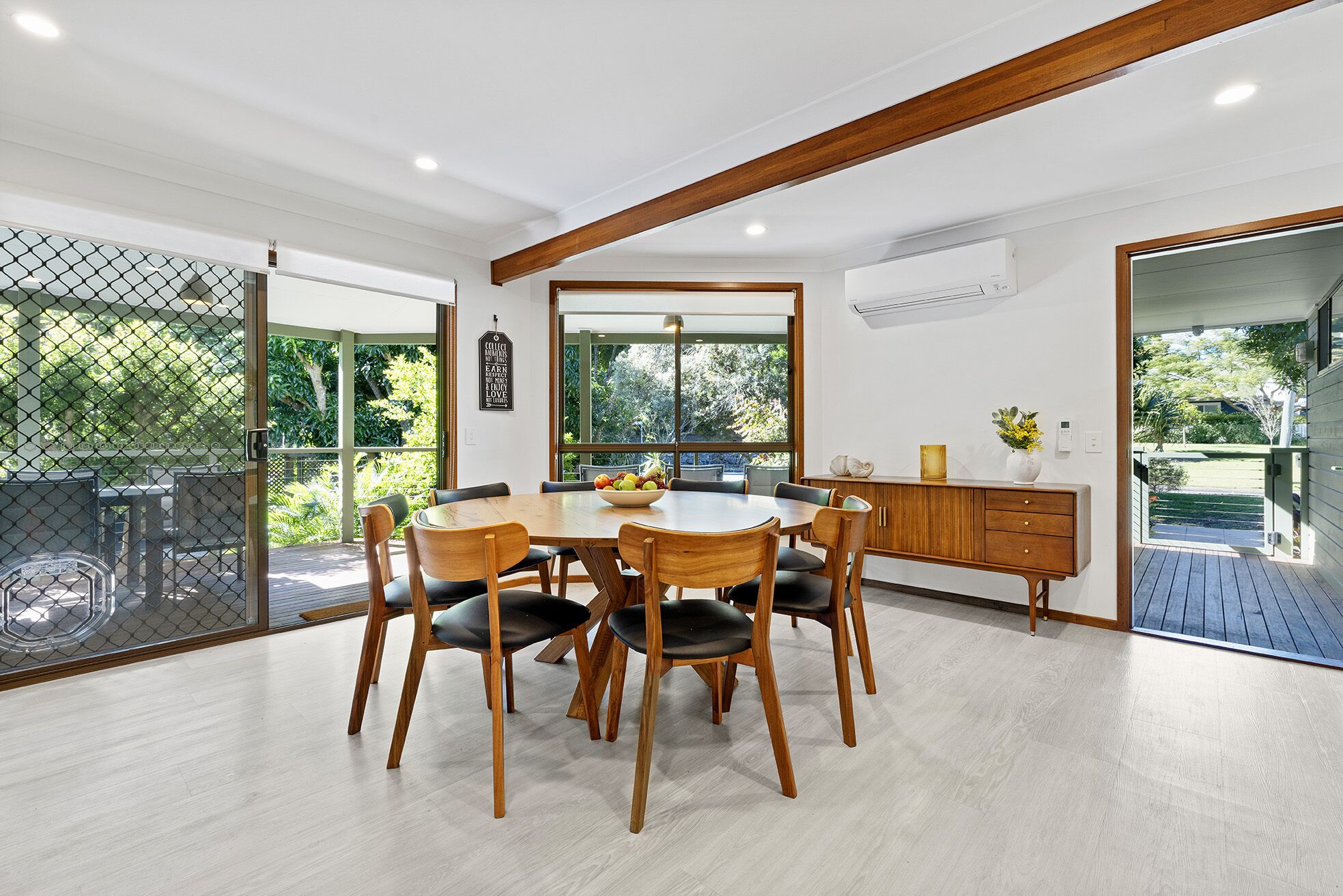 A dining area with a wooden table and chairs, a large window with a view of greenery, and a wooden ceiling beam.