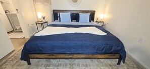 Un dormitorio con estructura de cama de madera, colcha azul, dos mesitas de noche con lámparas y una pared blanca.