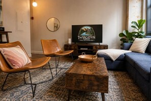 Ein Wohnzimmer mit Fernseher, zwei Ledersesseln, einer Couch und einem Couchtisch, sowie einer großen Pflanze.