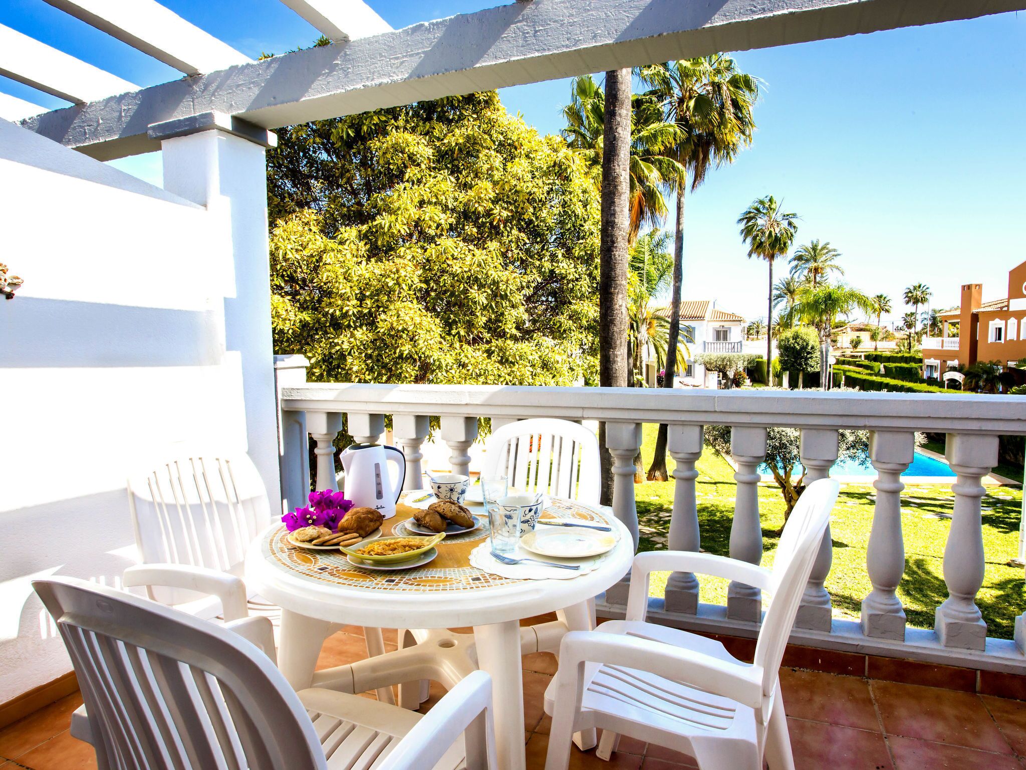A terrace with a white table set for dining, surrounded by white chairs, overlooking a pool and palm trees.