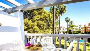 Una terraza con mesa blanca lista para comer, rodeada de sillas blancas, con vista a una piscina y palmeras.