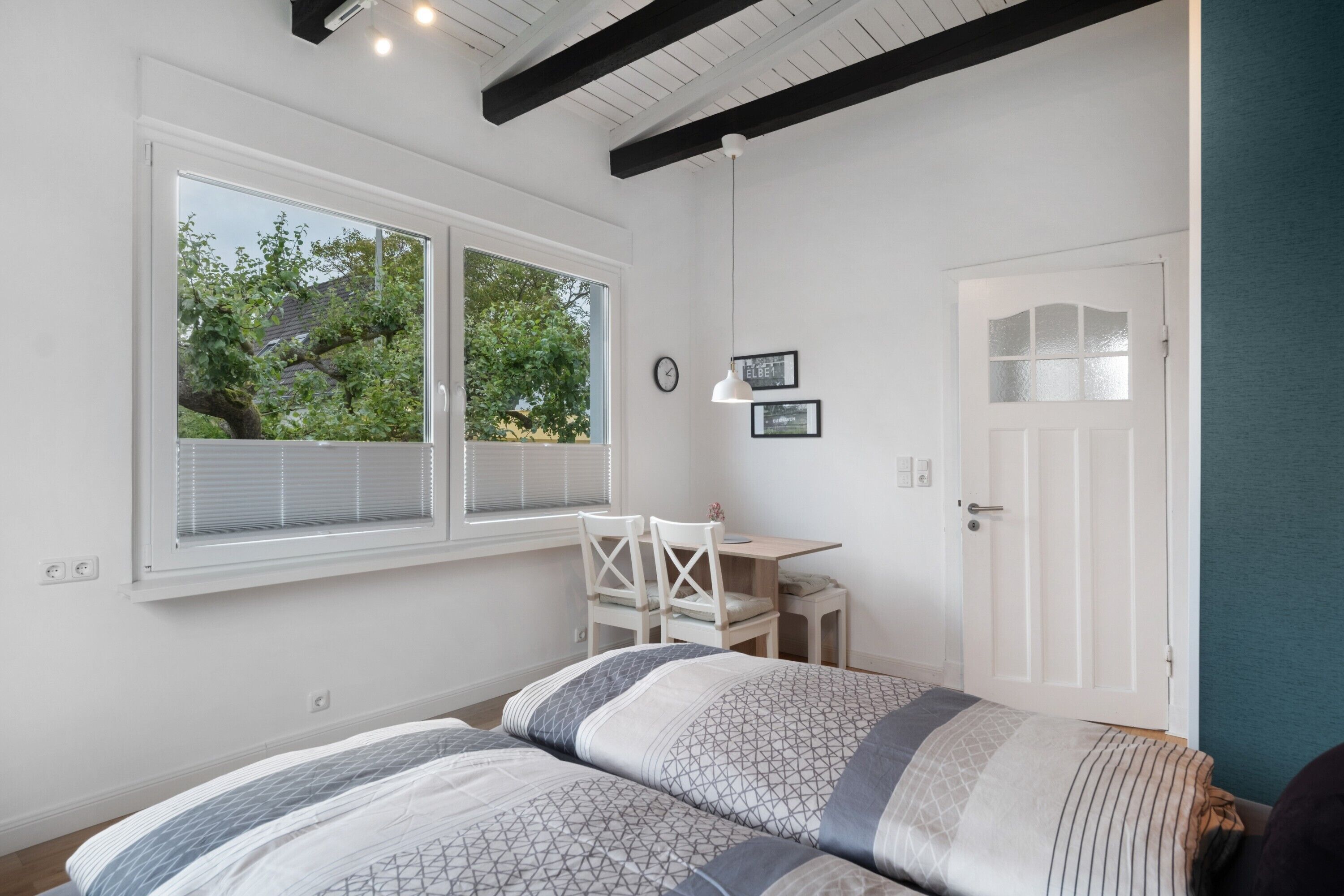 A bedroom with a bed, a desk, and a chair. There is a window with blinds, a door, and a clock on the wall.