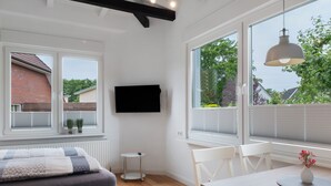 Una habitación moderna con cama, una mesa de comedor con sillas, un televisor montado en la pared y ventanas amplias con vistas al exterior.