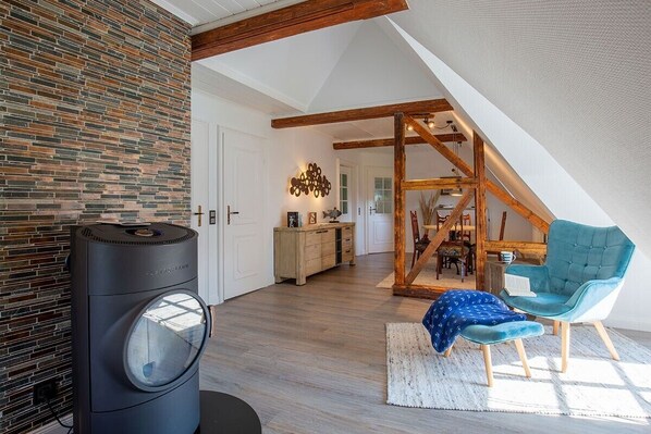 Une pièce de style « loft », avec des poutres en bois apparentes, un mur en pierre, un radiateur noir, une chaise bleue et un escalier menant à un niveau supérieur.