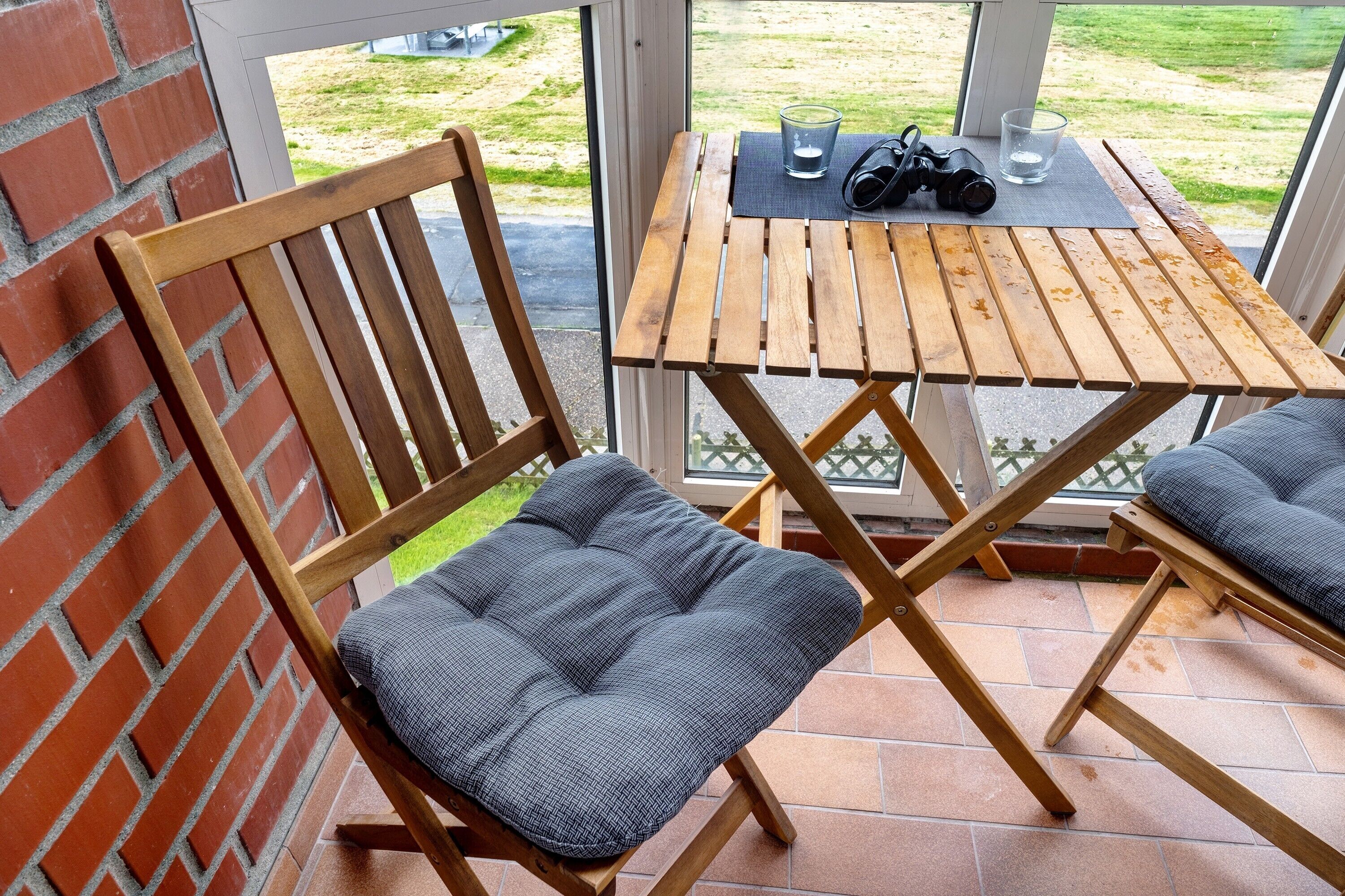 Een houten stoel met een bekleed zitvlak en rugleuning, een houten tafel met een glas en camera erop, en uitzicht op een grasveld door een glazen deur.