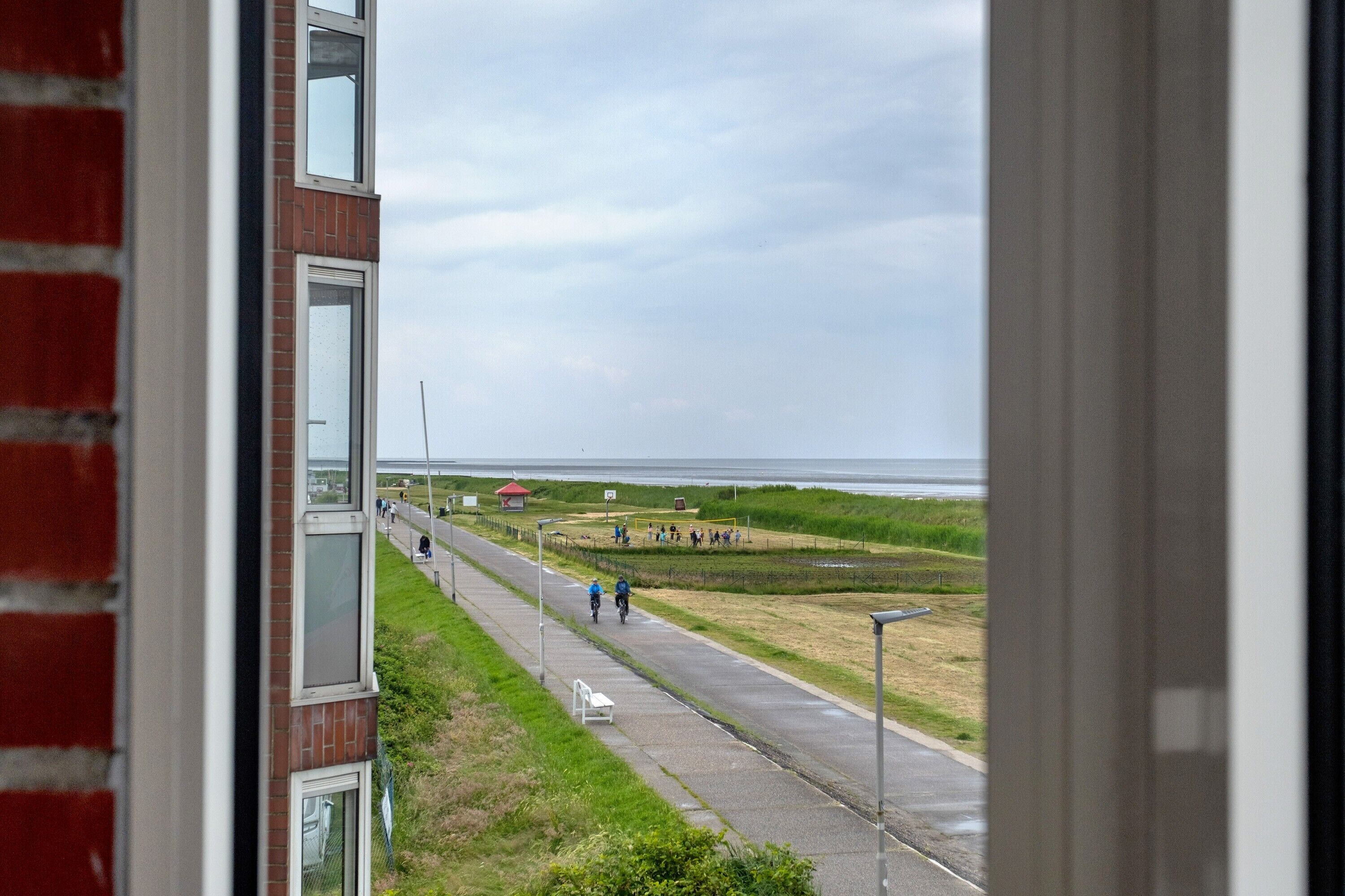 A view from a window overlooking a coastal road with a bench, a brick building, and a distant body of water.