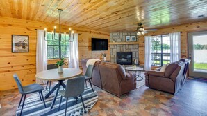 Interior de una cabaña de madera con chimenea de piedra, un sofá, una mesa de comedor con sillas y un televisor montado en la pared.