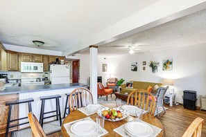 Une cuisine moderne avec des meubles en bois et un îlot central, un coin repas avec une table en bois pour quatre personnes, et un salon avec un canapé et des fauteuils.