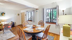 Un coin repas avec une table ronde dressée pour quatre personnes, entourée de chaises en bois. On y trouve une fenêtre avec des rideaux, une lampe et un tableau accroché au mur.
