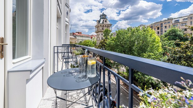 A balcony with a table and chairs, a view of a historic building with a clock tower, and lush greenery. - Apartment in Clouds Letná Luxury Balcony (Prague)