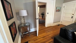Un salón con piso de madera, un sofá de cuero negro, una mesita de noche con una lámpara y una puerta que da a otra habitación.