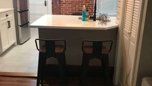 A kitchen with a small island, two bar stools, a refrigerator, and a window with blinds.