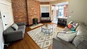 A living room with a brick wall, a fireplace, a television, and a dining area with chairs.