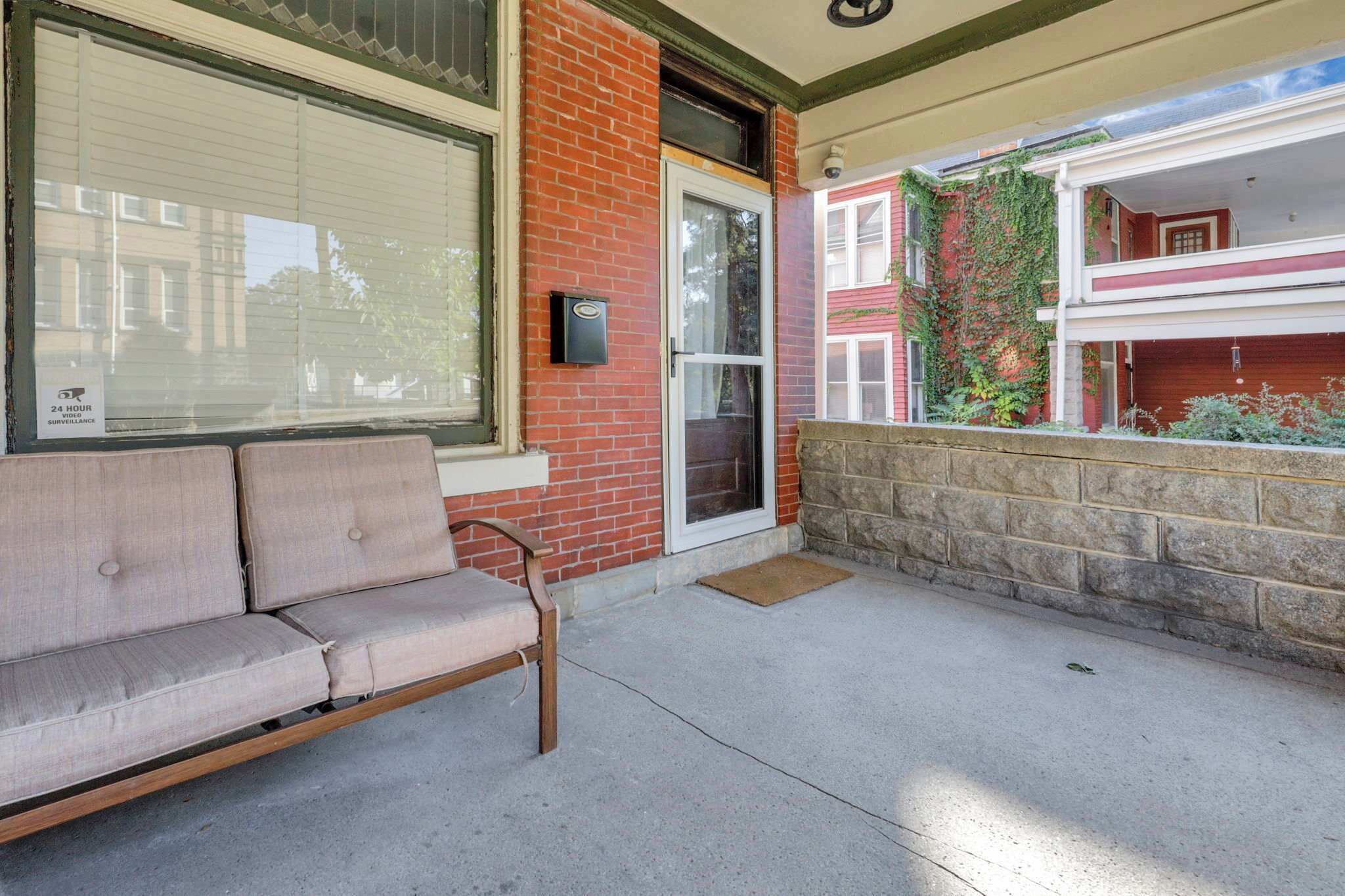A covered outdoor seating area with a bench, a brick wall, and a window with blinds.