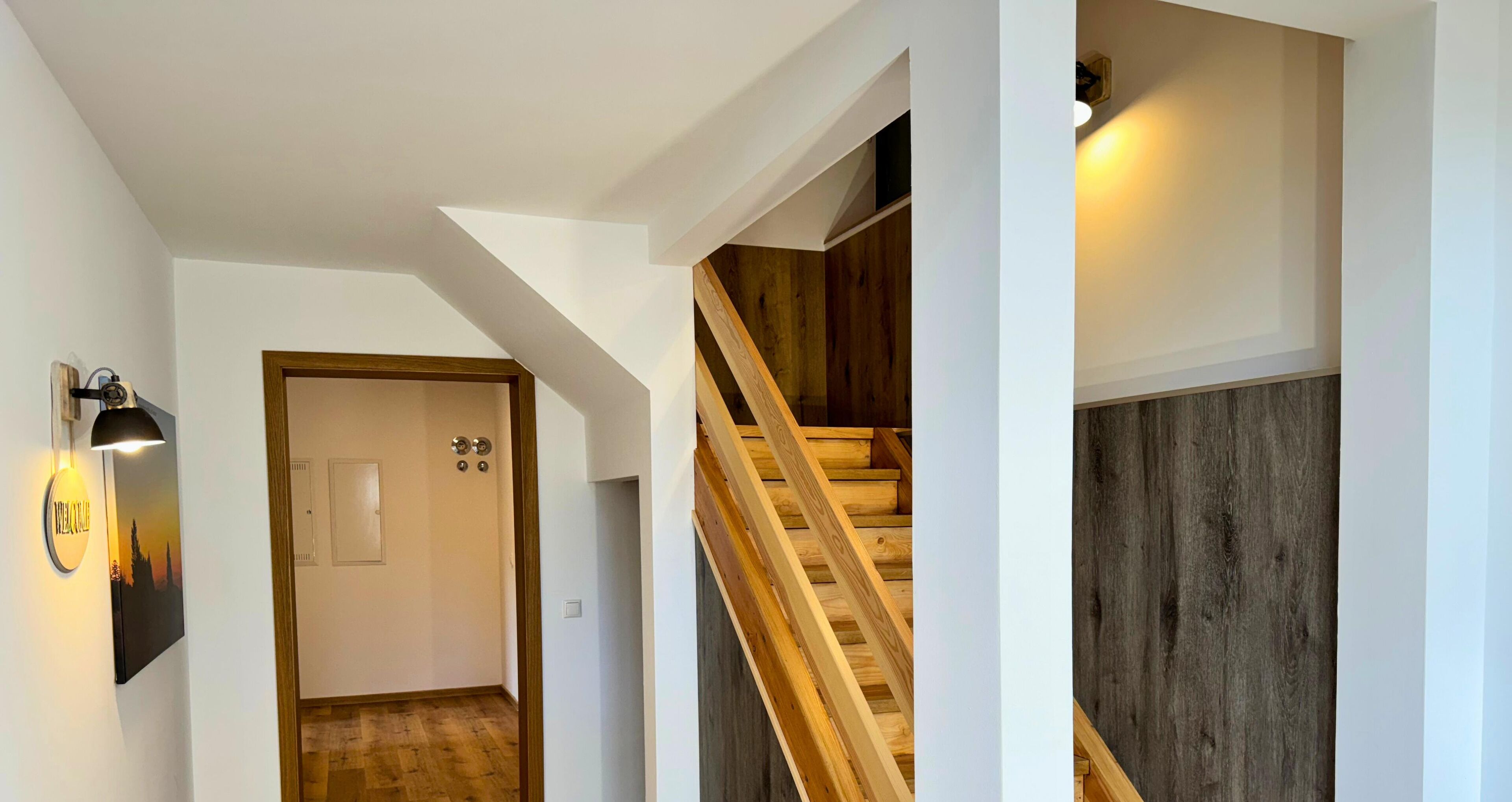 Un couloir avec un escalier en bois, une porte dotée d’un interrupteur et un luminaire fixé au mur.