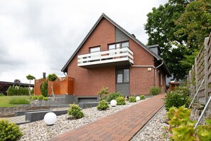 Une maison moderne avec une façade en briques, un balcon et un chemin bordé de lampes sphériques et de plantes.