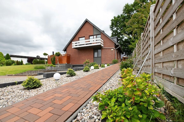 Une maison moderne avec une façade en briques, un balcon et un chemin bordé de gravier et de plantes.