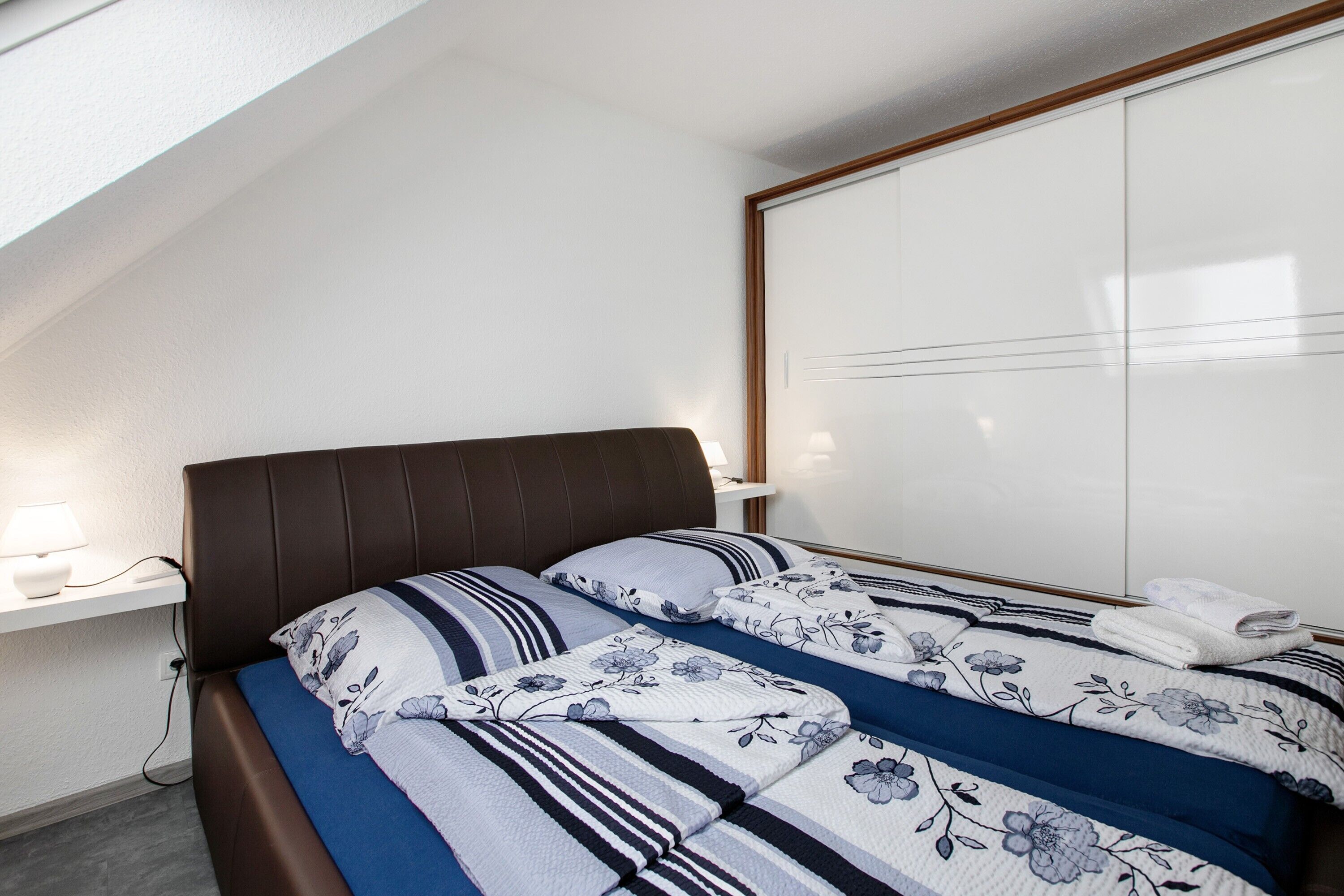 A bedroom with a bed, a nightstand, a wall-mounted shelf, and a wardrobe.