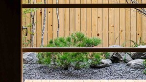 Vista a través de una ventana que muestra un cerdo de madera, un pequeño jardín con rocas y plantas, y un banco de madera en el interior.
