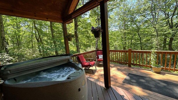 Un jacuzzi en una terraza de madera con sillas y barandilla con vistas a un bosque.