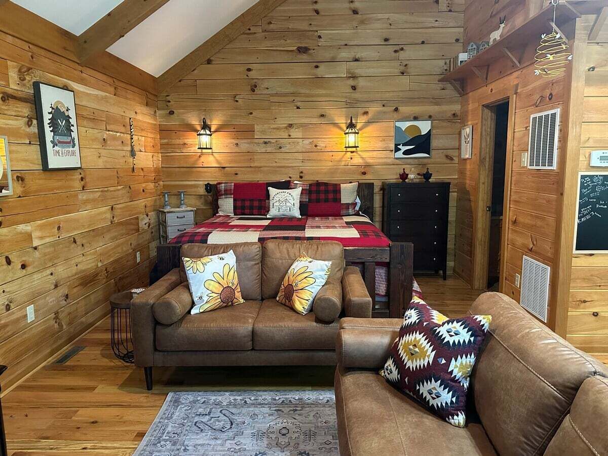L’intérieur d’un chalet confortable, avec un plafond en bois, un lit recouvert d’une couverture à carreaux rouges et noirs, deux canapés ornés de coussins à motifs floraux et des lanternes fixées au mur.