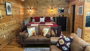 Un acogedor interior de cabaña de madera con techo de madera, una cama con manta a cuadros rojo y negro, dos sofás con cojines florales y faroles montados en la pared.