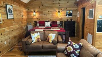 A cozy log cabin interior with a wooden ceiling, a bed with a red and black checkered blanket, two sofas with floral cushions, and wall-mounted lanterns.
