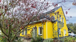 Uma casa amarela com escadas, um banco e uma árvore em primeiro plano.